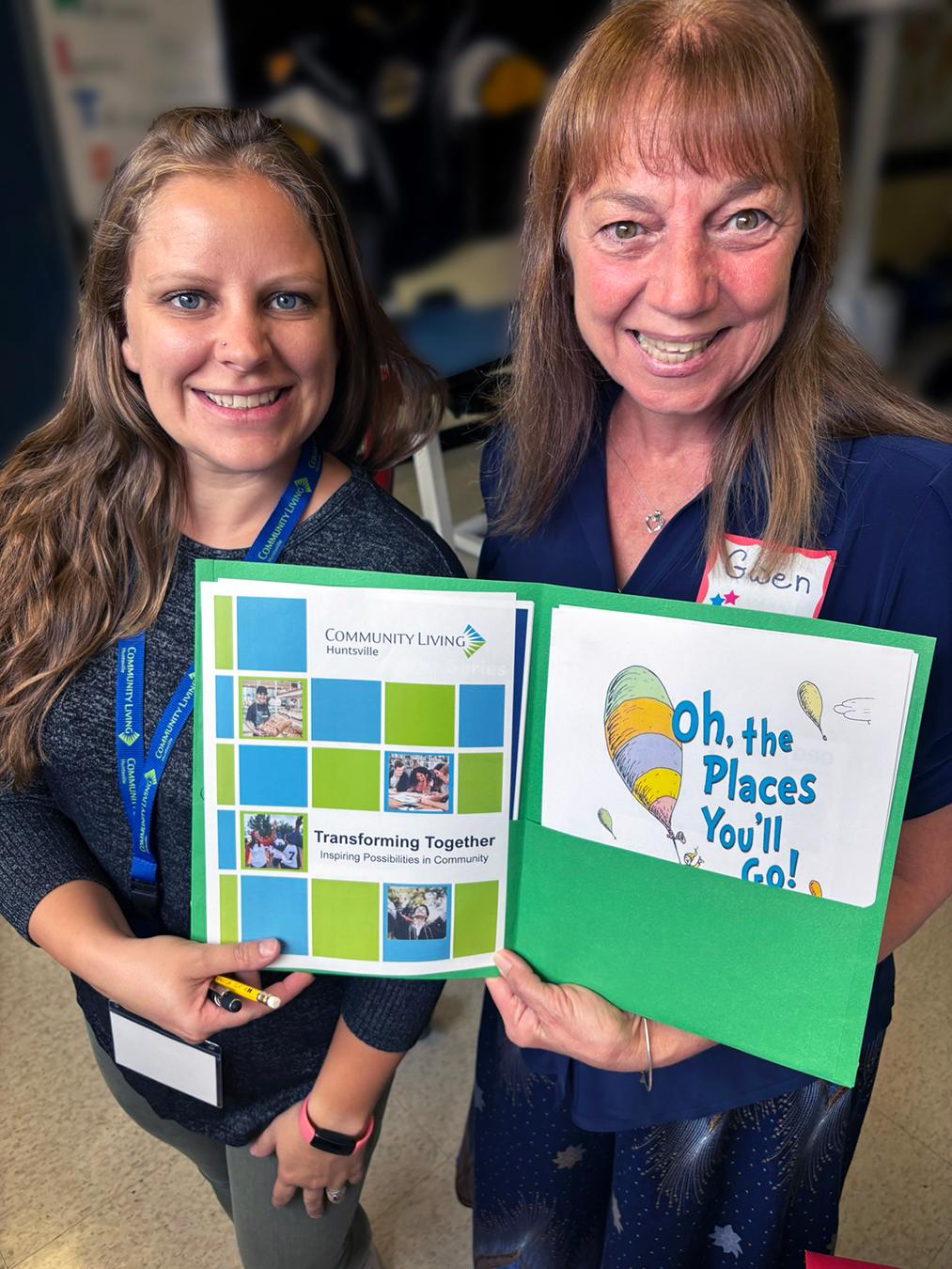2 smiling women in a classroom holding a folder with a front page that reads Transforming Together