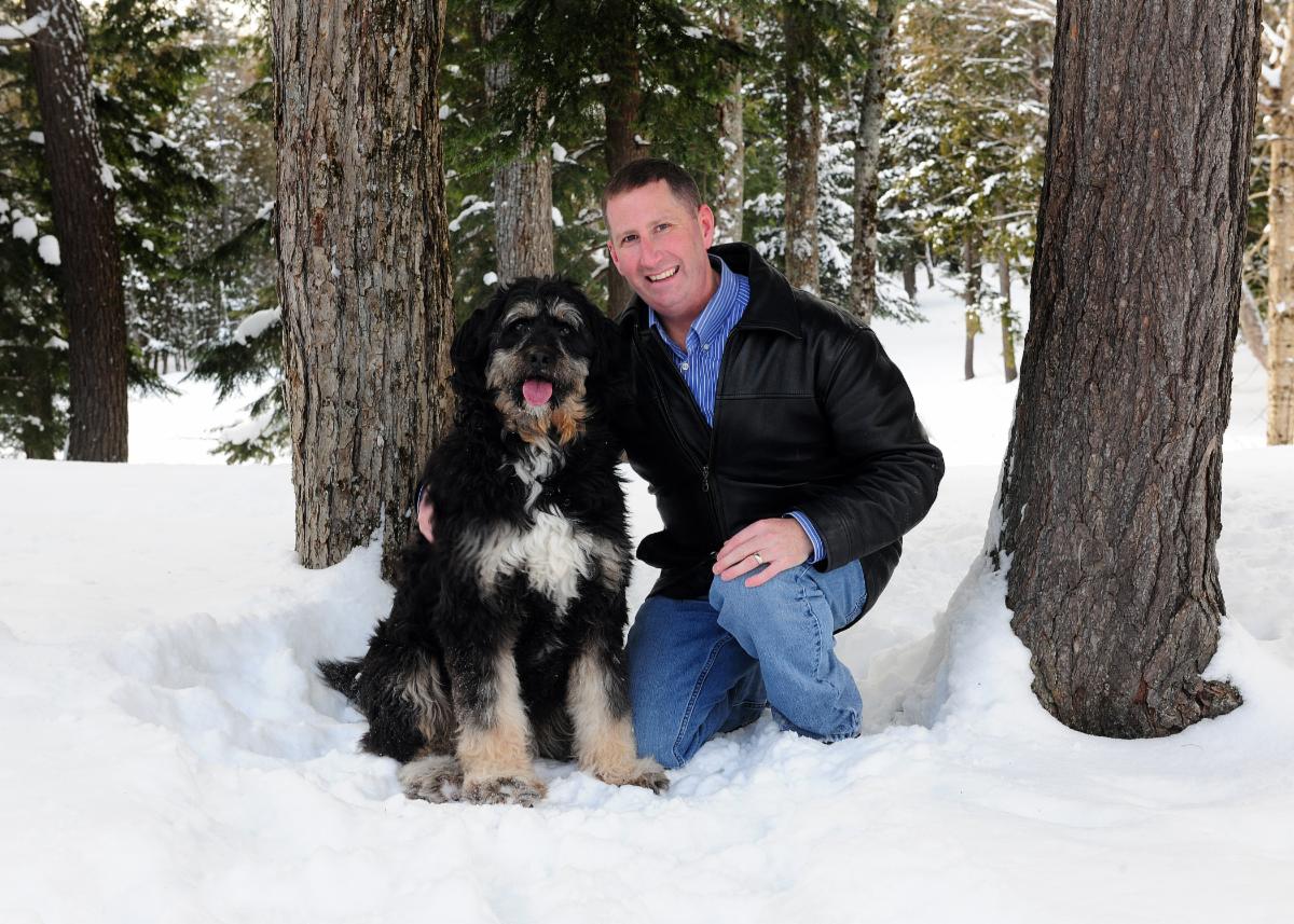 A smiling man crouching by a medium sized black and white dog in the snowy woods
