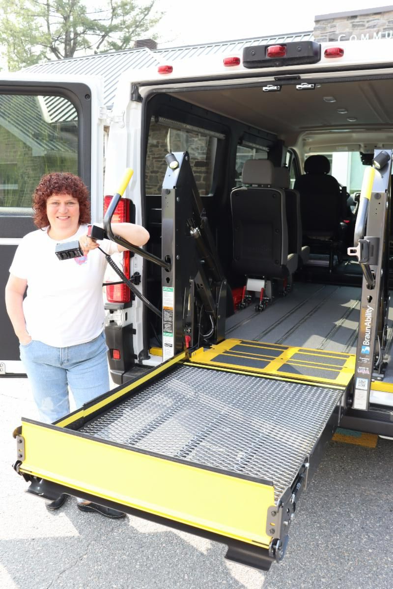 A smiling woman stands beside a raised wheelchair lift that comes out the back door of a white accessible van
