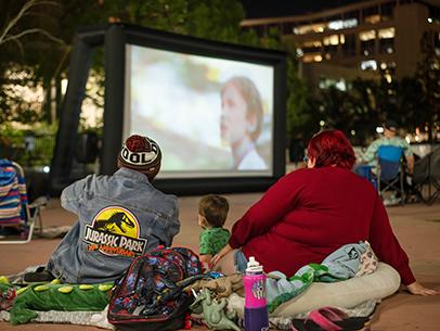 an adult sits and watches a movie on a big screen outside with a child and another adult.