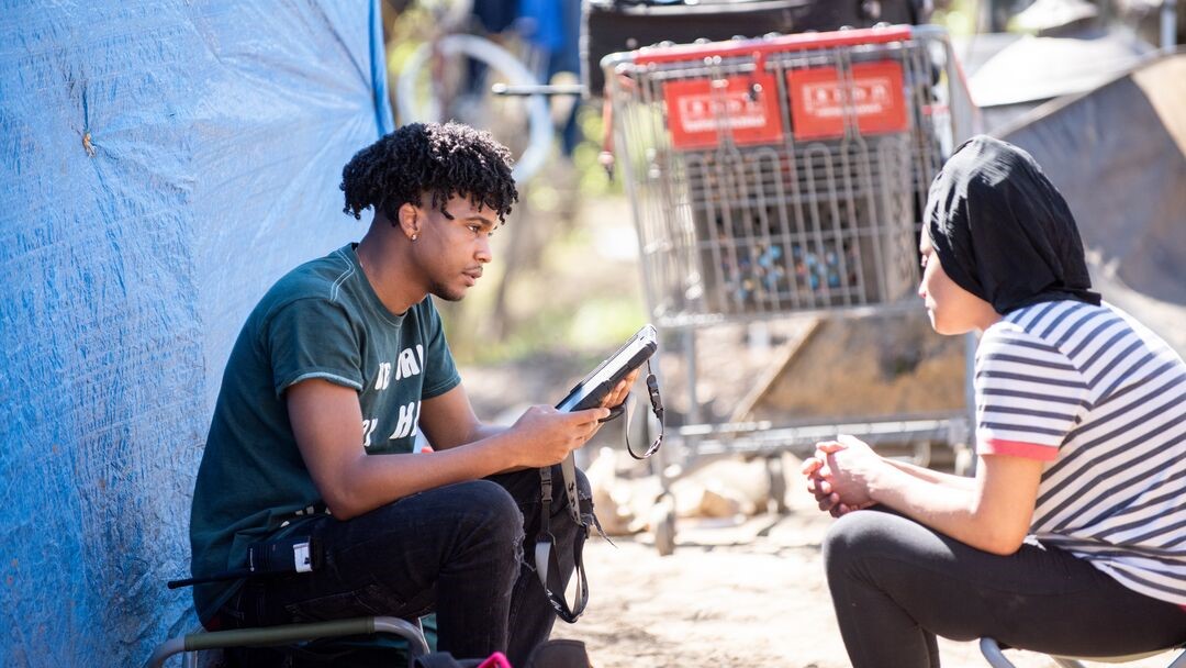 Man Outside Tent Holding Clipboard_ Talking to Seated Woman Facing Him.jpg
