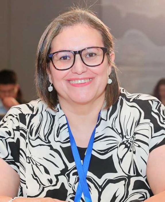 ICANN82 Fellow Rolla Hassan. She's smiling and wearing glasses, a black and white shirt, and a blue lanyard around her neck.