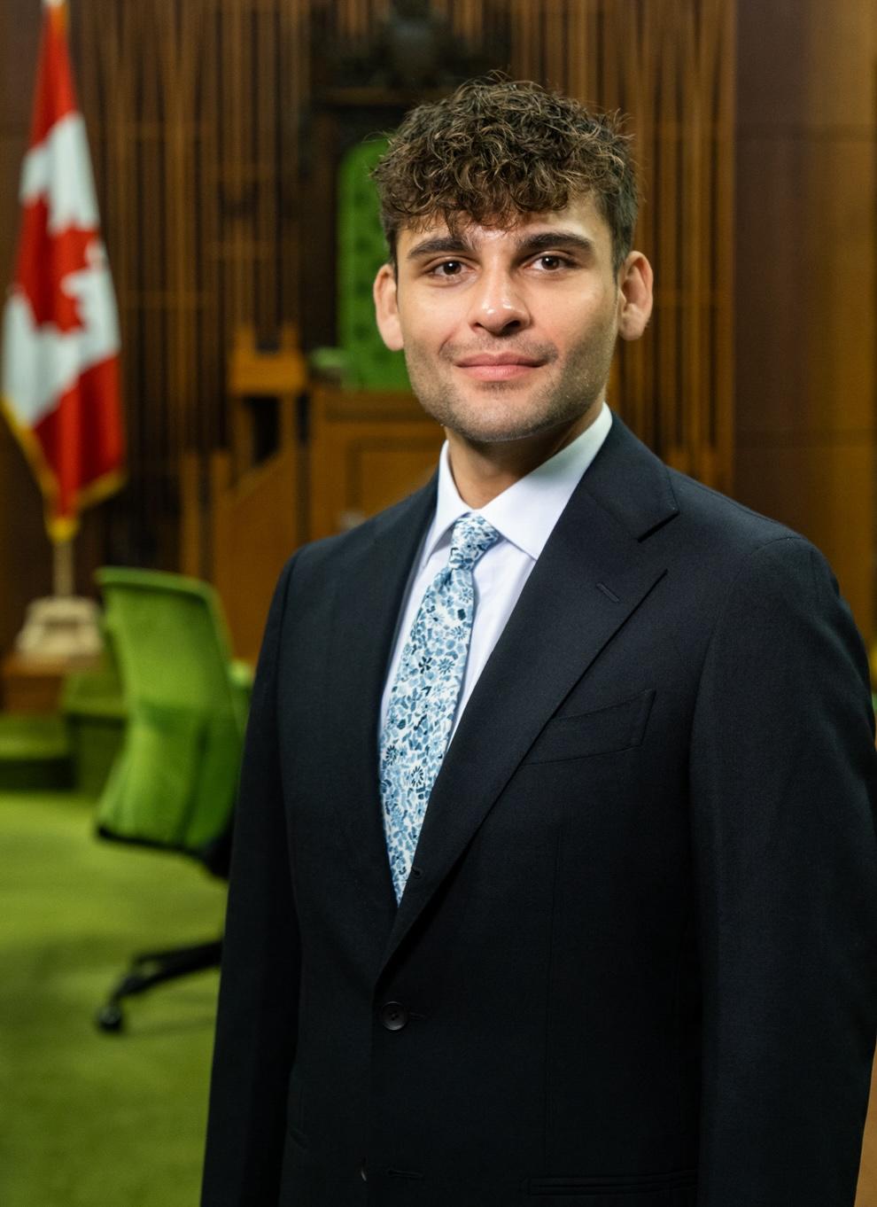 ICANN82 NextGen participant Justin Langan. He's wearing a dark blue suit with a light blue tie., and is standing in a Canadian government chamber.
