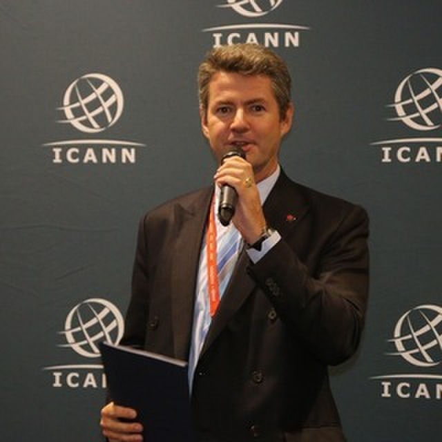 Olivier Crépin-Leblond, Co-Chair, At-Large Consolidated Policy Working Group (CPWG). He's speaking into a microphone while standing in front of an ICANN-branded background. He's wearing a navy suit, light blue striped tie, and orange lanyard around his neck. 