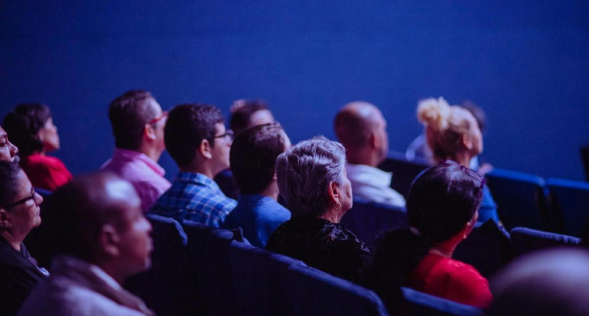 Photo of a crowd in a theater, looking up at a presentation. We’re facing their backs.