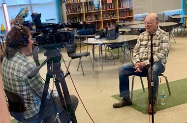 Photo of person in a chair talking to a videographer next to camera.