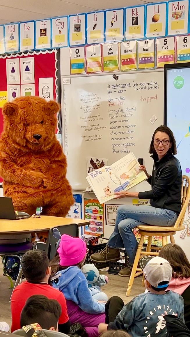 Mayor Perri Melnick reading a book to children in a classroom with a grizzly bear mascot sitting to her left.