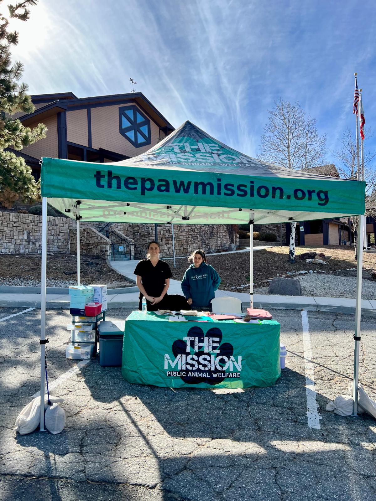Image of two women standing under a green tent with The Paw Mission logo on it in front of Big Bear Lake City Hall.