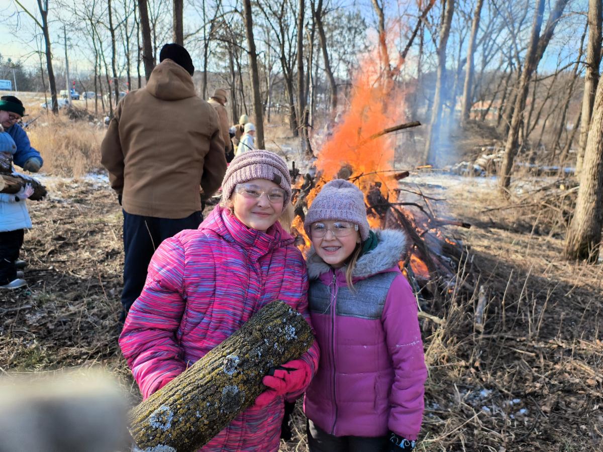 2 students in woods with bonfire behind them