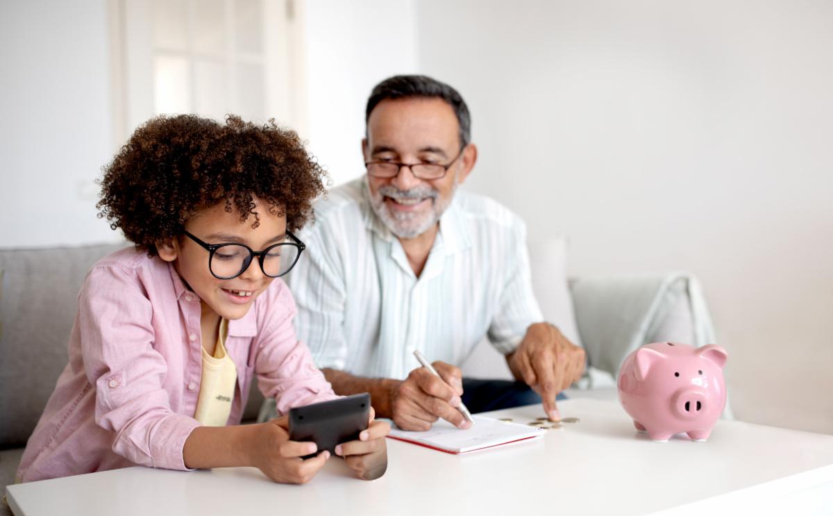 Money Skills. Smiling Senior Grandpa and Grandson Boy Engaging in Finance Education Hold Wallet And Calculating Savings with Calculator Sitting Near Table With Piggybank At Home. Selective Focus