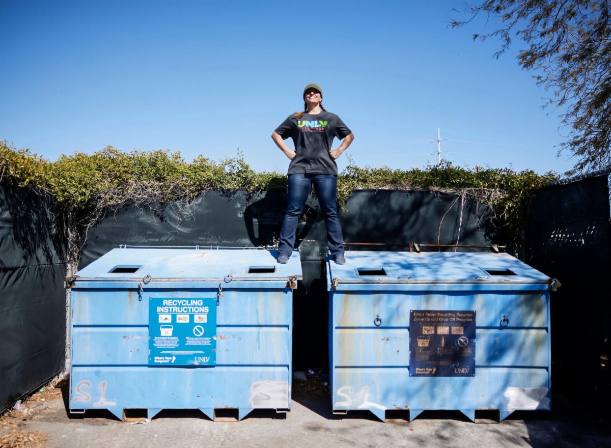 A smiling Tara Pike stands on two blue recycling dumpsters.