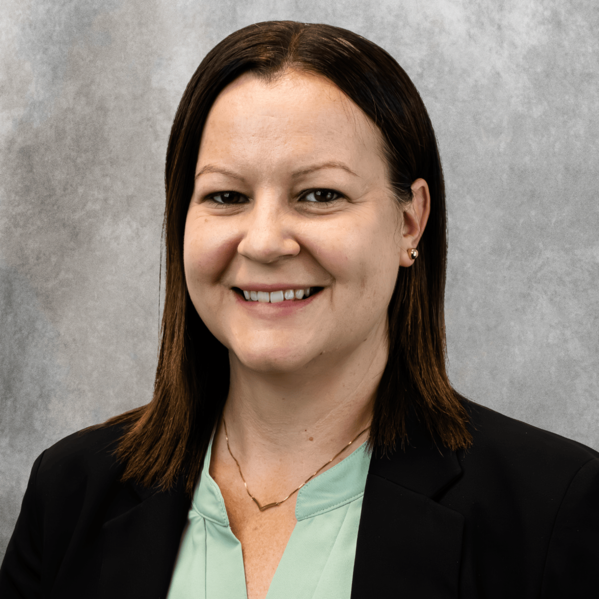 Laura Schneider from Quality Assurance & Financial Compliance posing in front of a gray background with a light green shirt and black blazer 