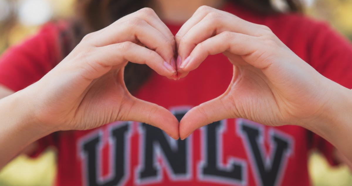 A person holding up heart hands in the foreground with a red UNLV shirt in the background