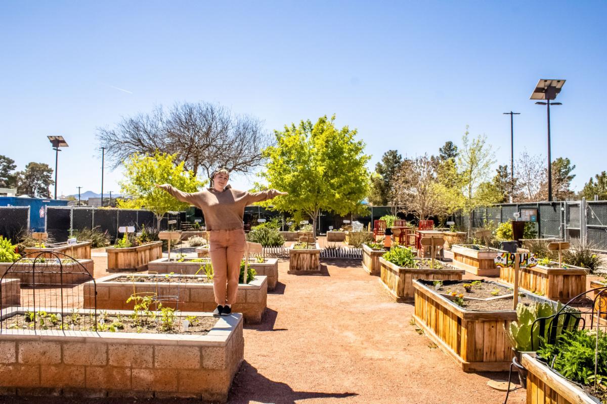 Andie stands on a garden bed platform at the Community Garden with her hands outstretched