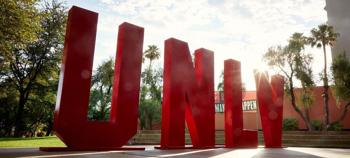 This is a picture of red UNLV letters against a blue sky and clouds with trees in the background.