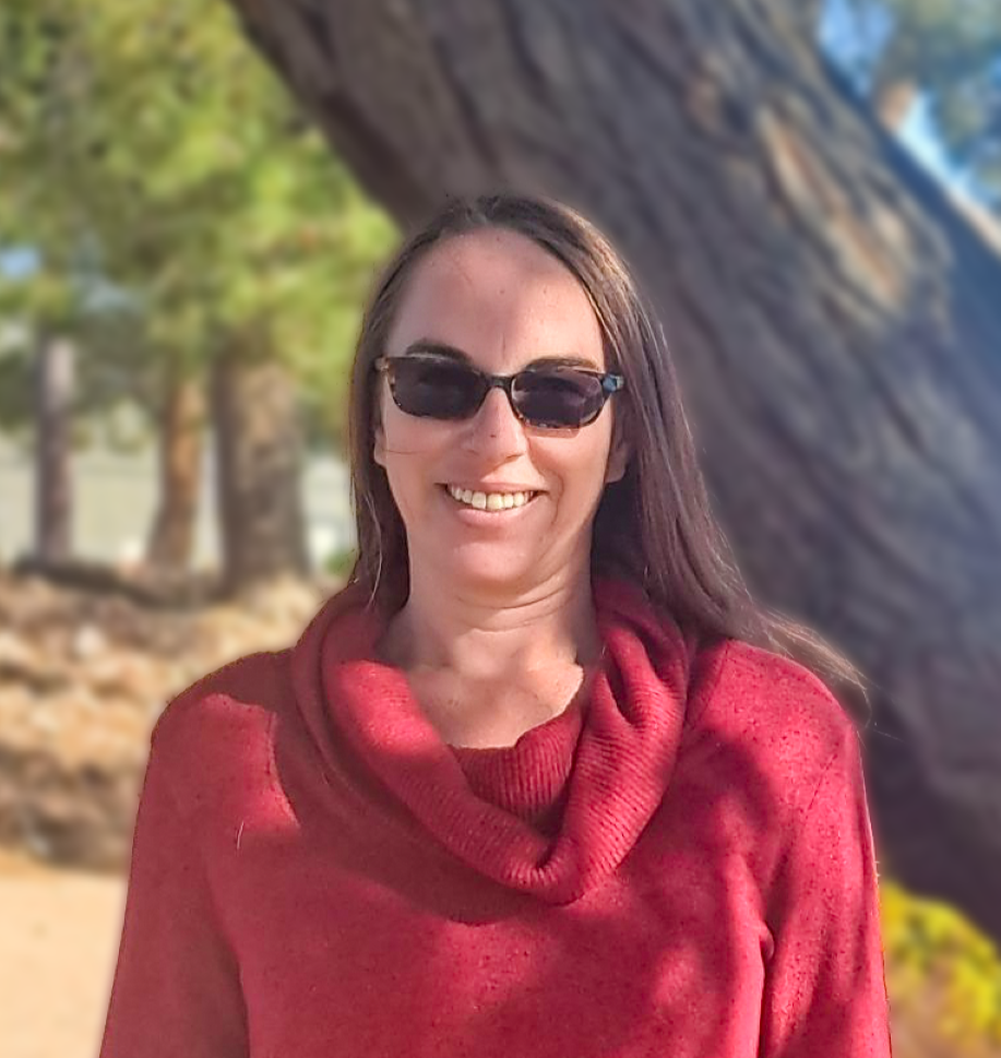 A white woman in front of a tree with long brown hair dark sunglasses and a red cowlneck sweater on