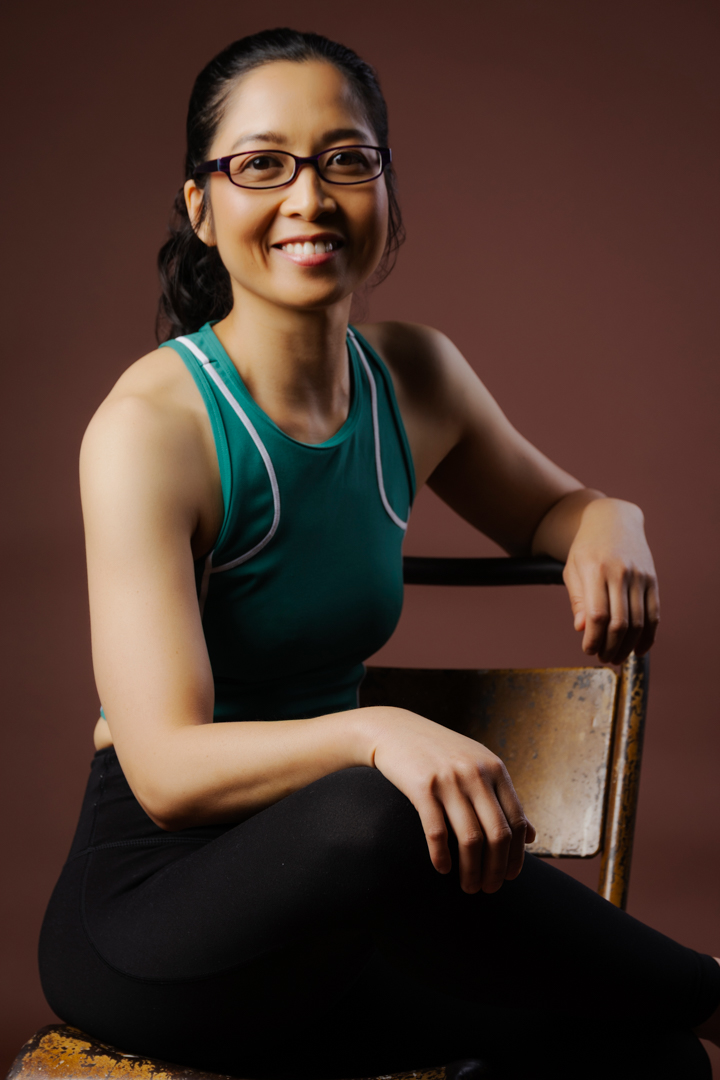 A Filipino woman wearing glasses, a green singlet and black pands is sitting on a chair and smiling.