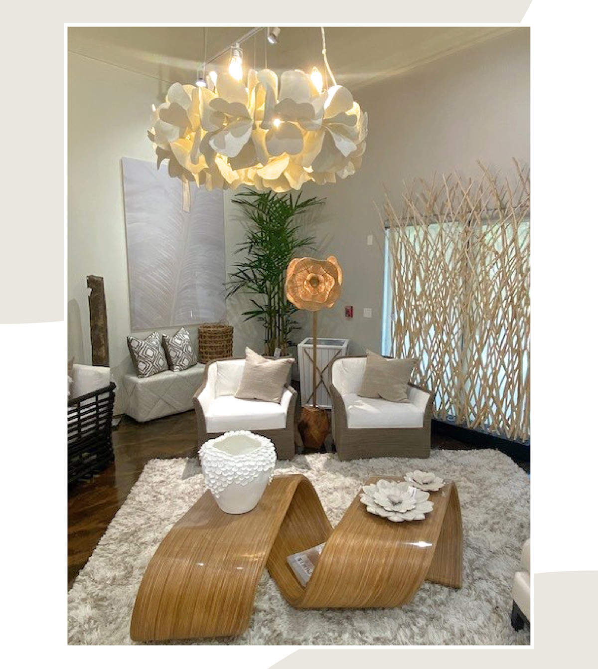 Palecek showroom vignette with a sculptural white flower-petal pendant light above two wicker lounge chairs with white cushions, a shag rug, and glossy bent-wood coffee tables topped with a textured white vase and ceramic flowers.