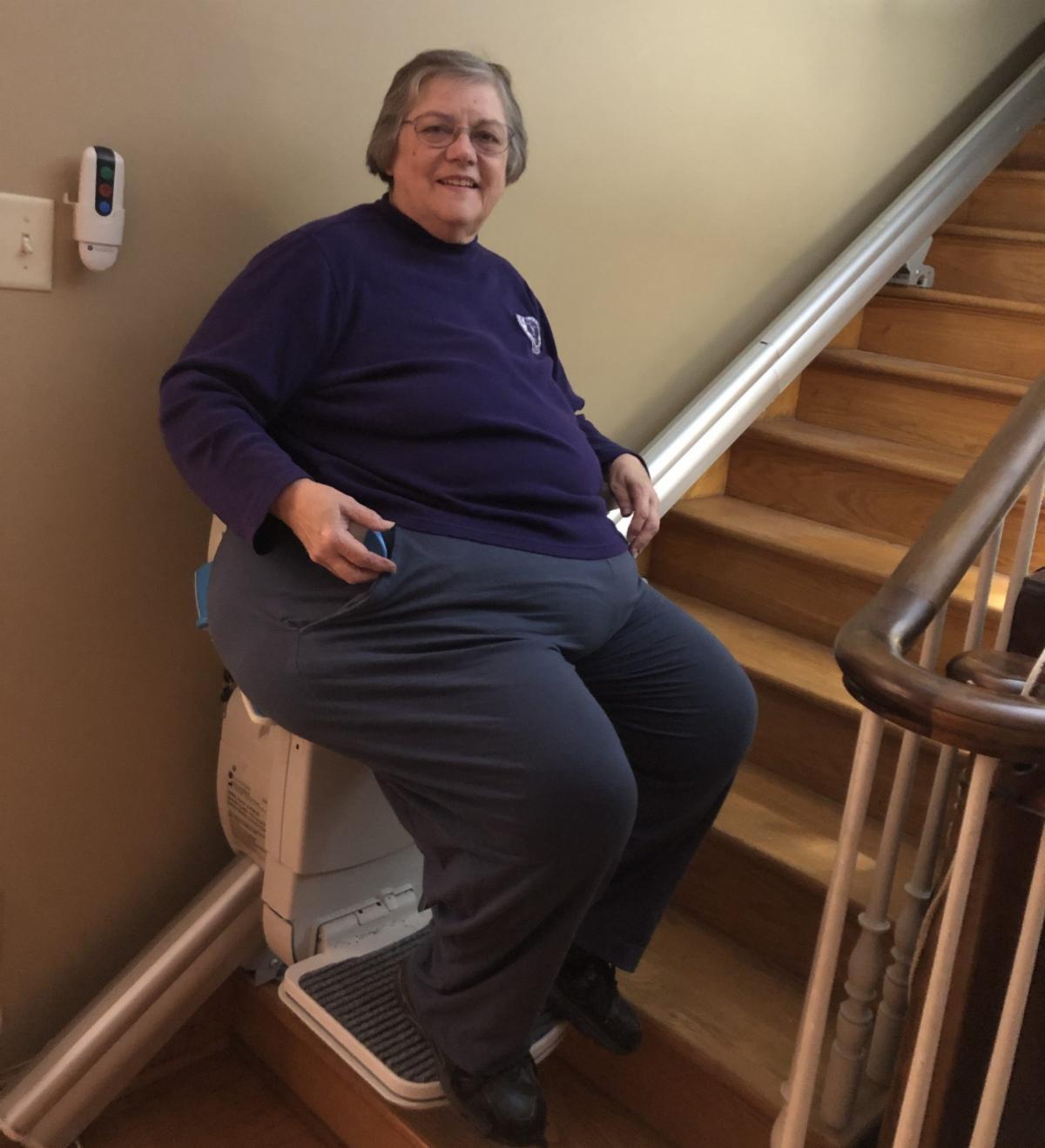 Cara Beth is a woman with short hair and glasses. She is wearing a blue long sleeve top and gray long pants. She is sitting on her chairlift on the stairs.