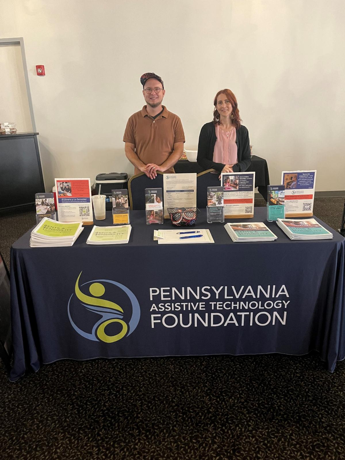 There is a man in a brown shirt wearing a baseball hat and a woman in a pink top and black cardigan standing behind a table that has resources on it and a table cloth that has Pennsylvania Assistive Technology Foundation on it.