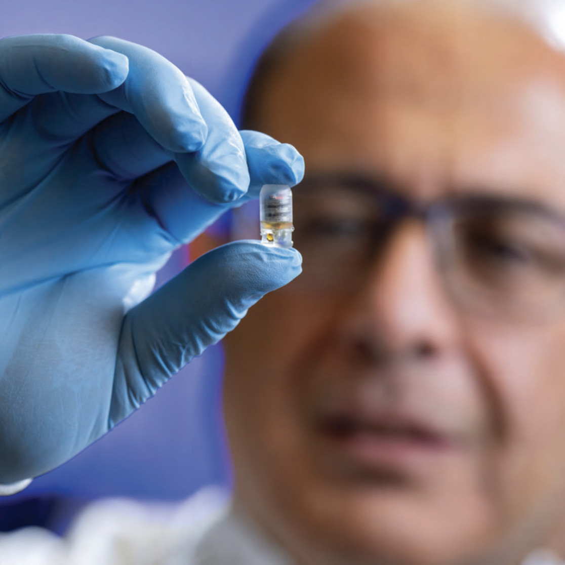 tiny capsule is in focus and held up in a blue lab glove by Reza, whose face is out of focus behind the pill