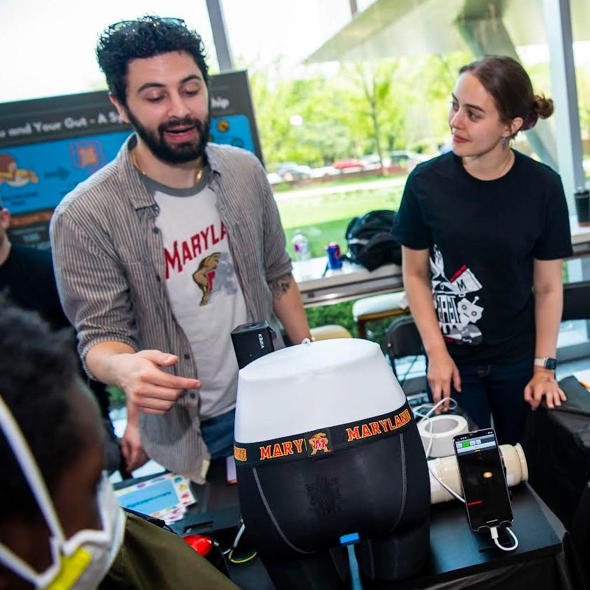 Two individuals at a science demonstration booth, discussing a model displaying anatomy. One man, wearing a "Maryland" shirt, gestures towards the model, while a woman in a black shirt listens attentively.