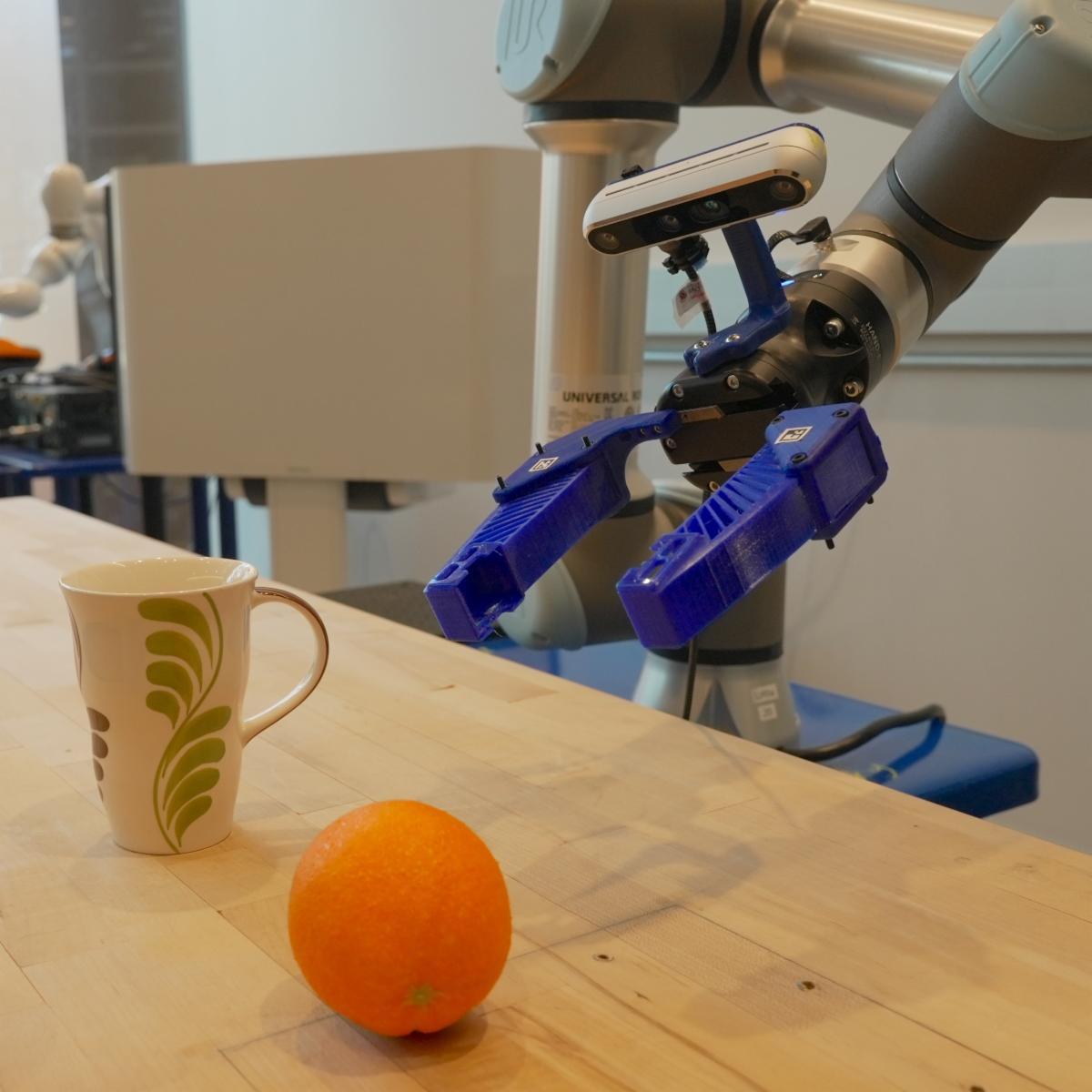 A robotic arm hovers above a mug and an orange on a table.
