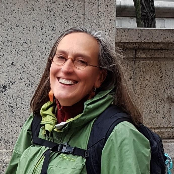 A woman with long brown hair wearing round glasses a green jacket and a backpack smiles at the camera.