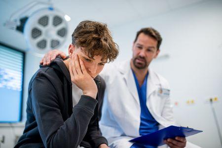 A doctor sits next to a distressed teen patient in a hospital bed.