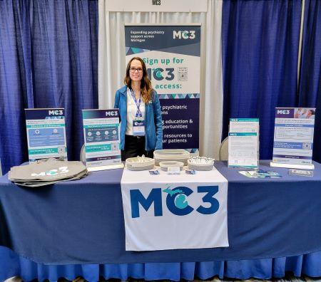 Jessica Jenkins standing behind an exhibit table with MC3 materials at an event.