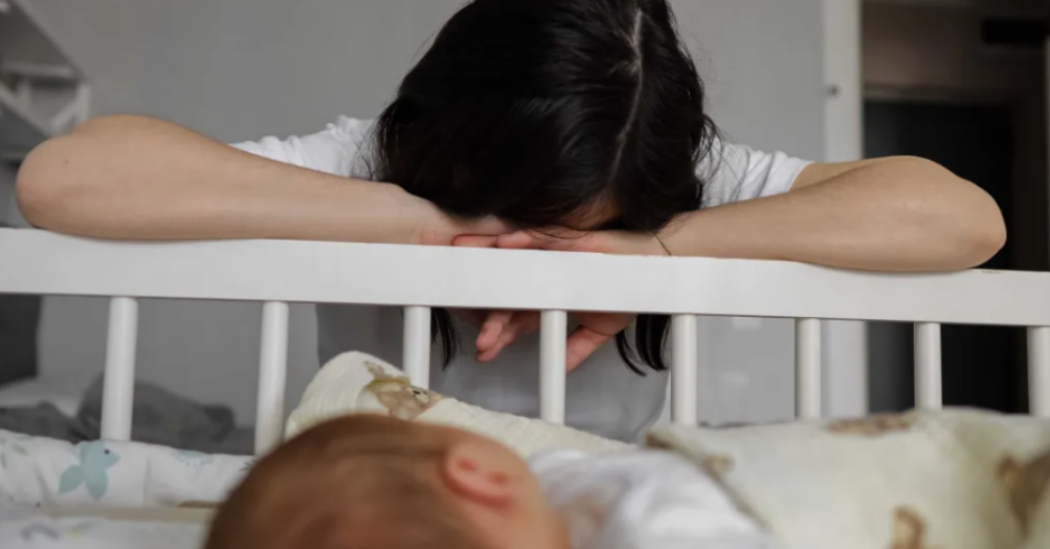 A mother rests her head on the side of a crib next to her sleeping baby.