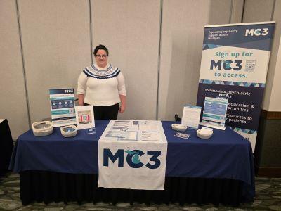 Sarah Wise standing behind an exhibit table with MC3 materials.