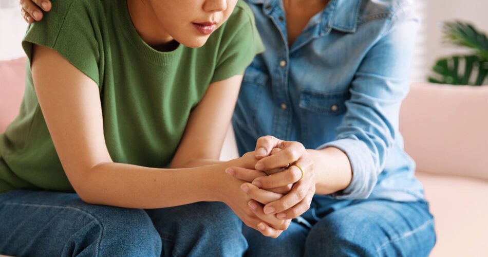 Distressed child sits on a couch next to a woman holding her hand.