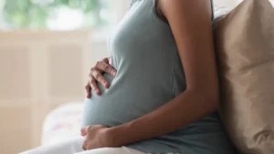 Pregnant woman sitting on a couch holding her stomach in front of a window.