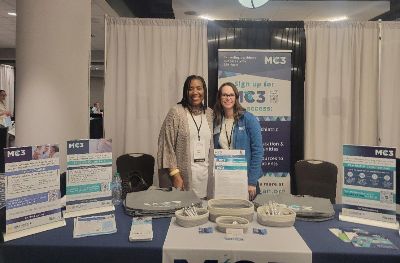 Jessica Jenkins and Carmen Harrison stand behind an exhibit table with MC3 materials at an event.