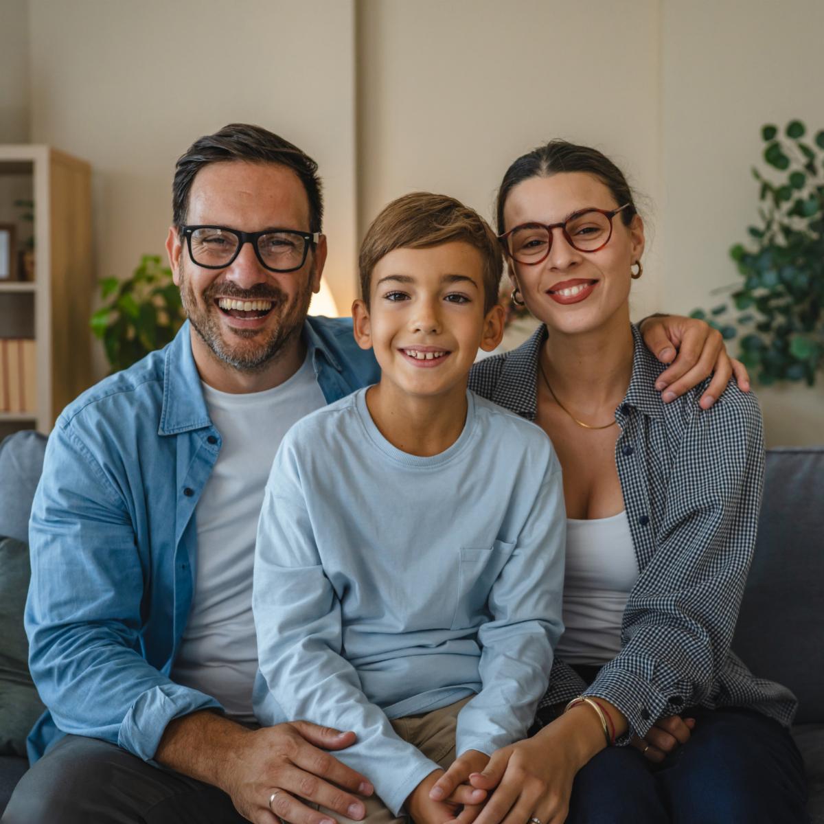 Smiling parents and young son wearing glasses pose together on a cozy sofa in their modern living room enjoying relaxed loving family time and warm togetherness at home