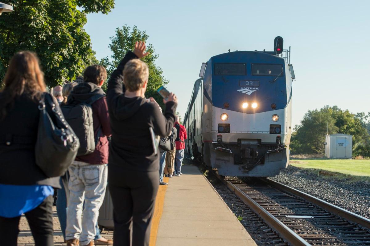 An Amtrak train approaches a Michigan station as passengers wait and wave from the station platform.