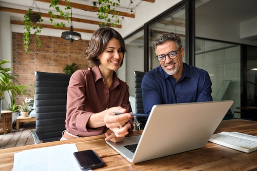 Business team of two executives working together using laptop in office. Happy young business woman manager showing project presentation to mature man client_ investor or partner at work meeting.