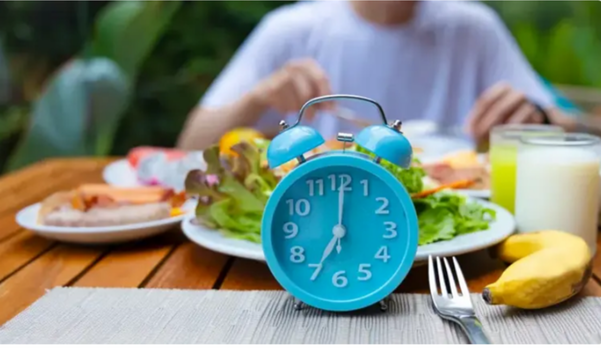 Little blue clock sitting on table, with plates of food behind it and person visible from the neck down, in white shirt, at the far end