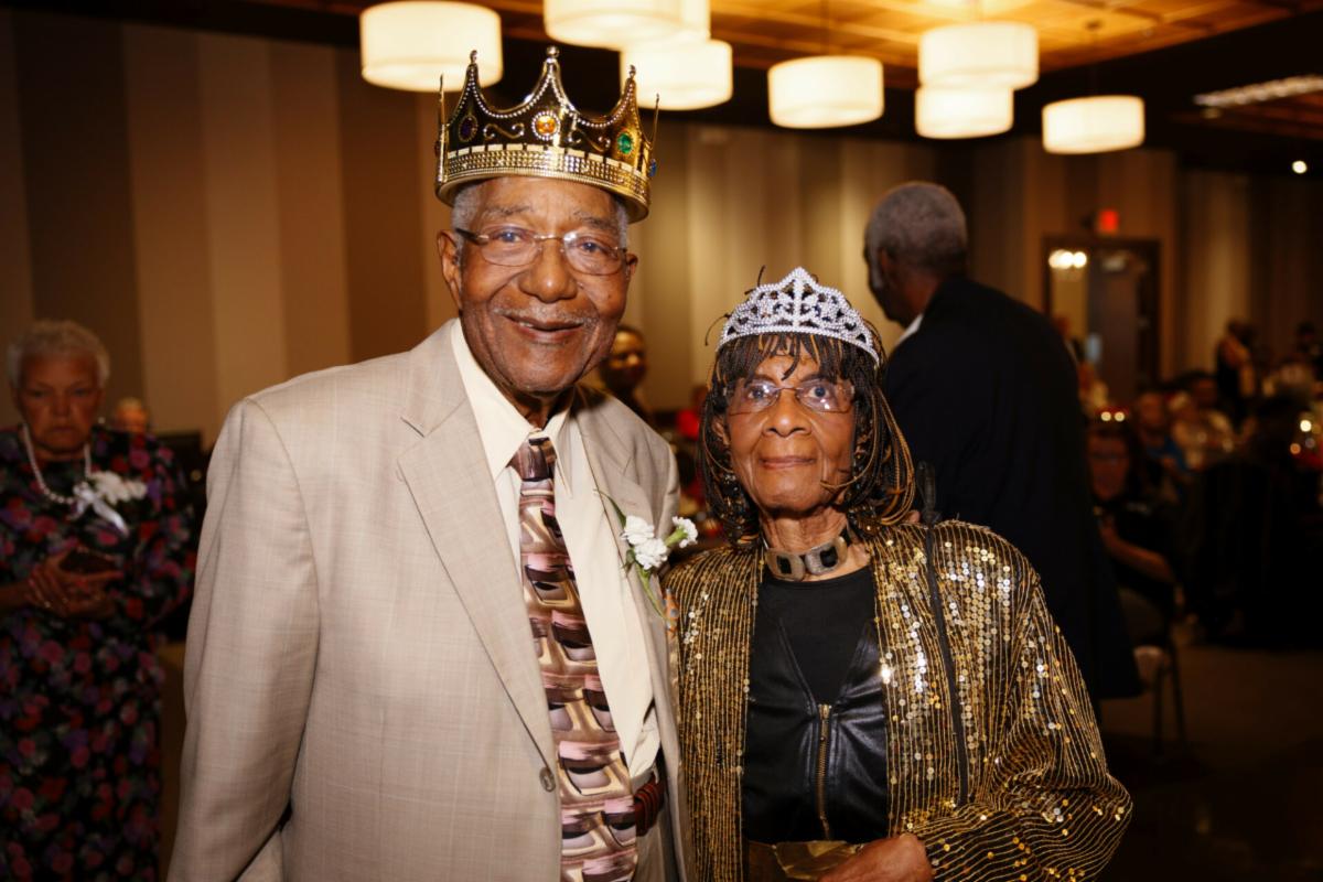 Older Black man wearing gold crown and older Black woman wearing silver crown at Senior Prom with others at tables in background