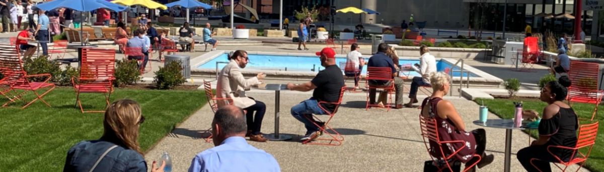 Banner showing people sitting around outdoor tables, some with blue or yellow table umbrellas, around an empty fountain in Downtown Toledo.