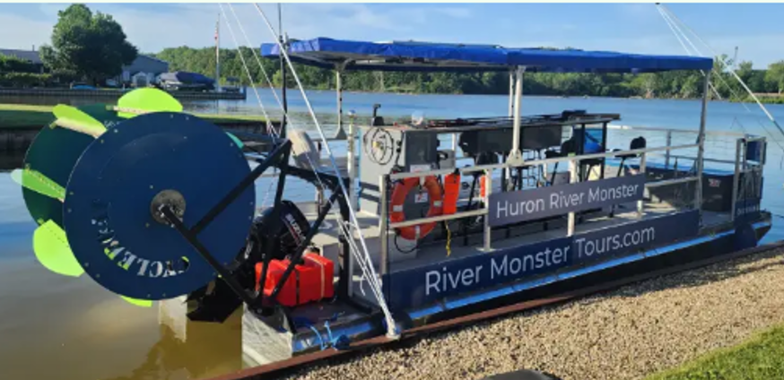 Boat named Huron River Monster with blue canopy and orange life jackets hanging on the side, docked with the Huron River in the background.