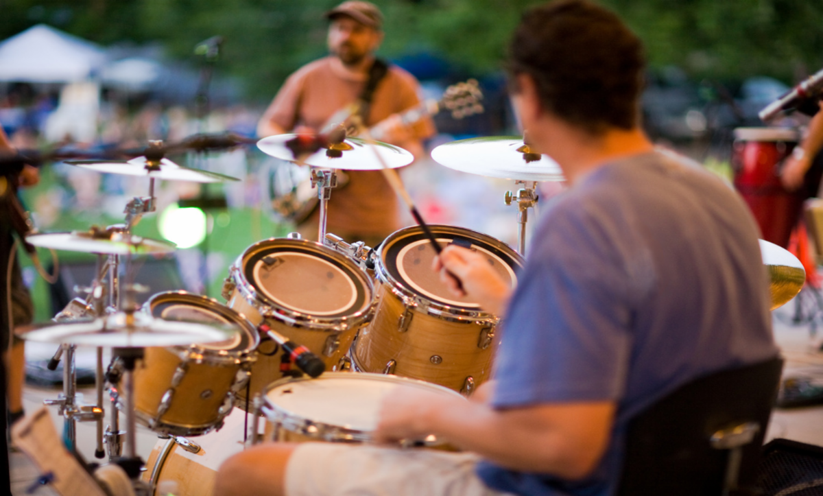 Close-up shot of musician with short hair behind drum and cymbal set, with man with guitar across the stage - and tents and tables in the distance