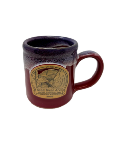 Limited edition 2026 Sand Dune Arch ceramic mug with a burgundy glaze, dark speckled rim, and an Arches National Park emblem featuring Sand Dune Arch.