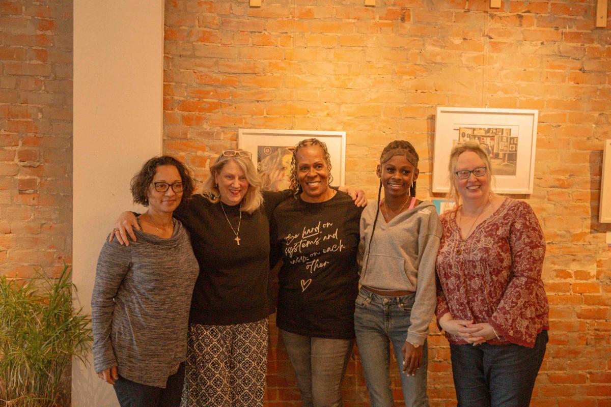 Five women standing together with their arms around each other in a warm group photo against an exposed brick wall. From left to right a woman with dark curly hair and glasses in a gray striped top a blonde woman in black with a cross necklace a woman in a dark sweatshirt with cursive text a young woman in a gray hoodie and jeans and a woman with glasses in a red patterned blouse. Framed artwork is visible on the brick wall behind them and there's a plant visible in the lower left corner.