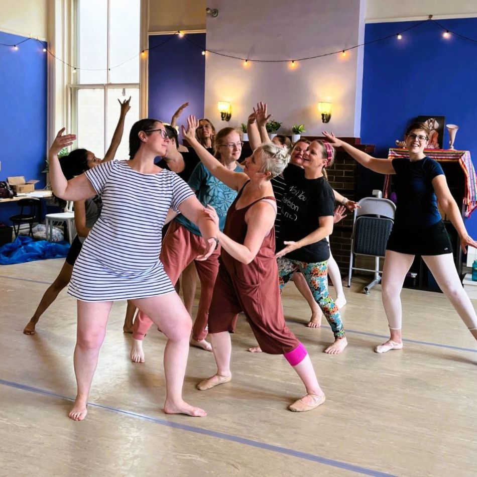 A group of dancers rehearsing in studio with arms raised a blue backdrop string lights hardwood floor