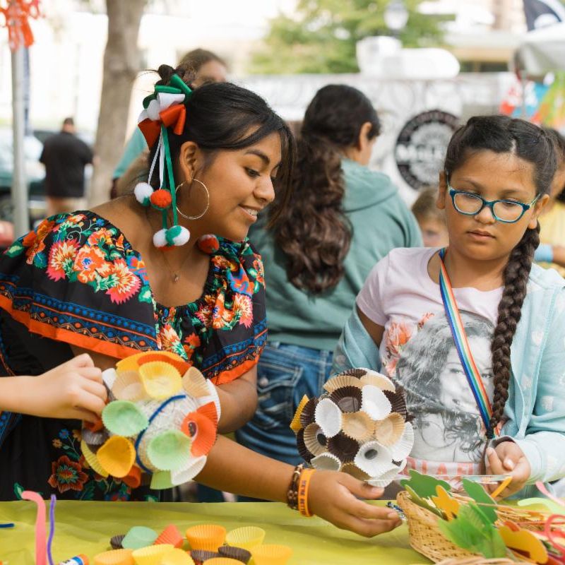 Two young people at an outdoor cultural festival or celebration sitting at a craft table making colorful paper plate decorations. The person on the left wears a vibrant floral top and has ribbons in the colors of the Mexican flag in their hair. They are holding up a paper plate decorated with bright circular cutouts. The person on the right wears glasses a light blue cardigan and has braided hair holding what appears to be a paper plate craft resembling an animal face. The table is covered with various colorful craft supplies including paper plates cutouts and other materials. Colorful decorative banners are visible in the background along with other festival attendees