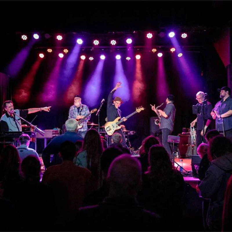 The image shows a live music performance in a small venue or club setting. A band of six musicians is performing on stage under dramatic purple, red, and blue stage lighting. The performers include musicians playing various instruments including guitar, bass, keyboards, saxophone, and percussion. Some of the band members appear to be gesturing or raising their hands while performing. In the foreground, the silhouettes of audience members can be seen watching the show from below the stage. The intimate venue has black curtains on the sides of the stage and professional lighting equipment mounted above. There's a photographer's watermark or logo visible in the bottom right corner of the image.