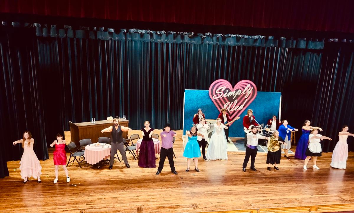 People dancing together in a line onstage, set in a wedding hall.