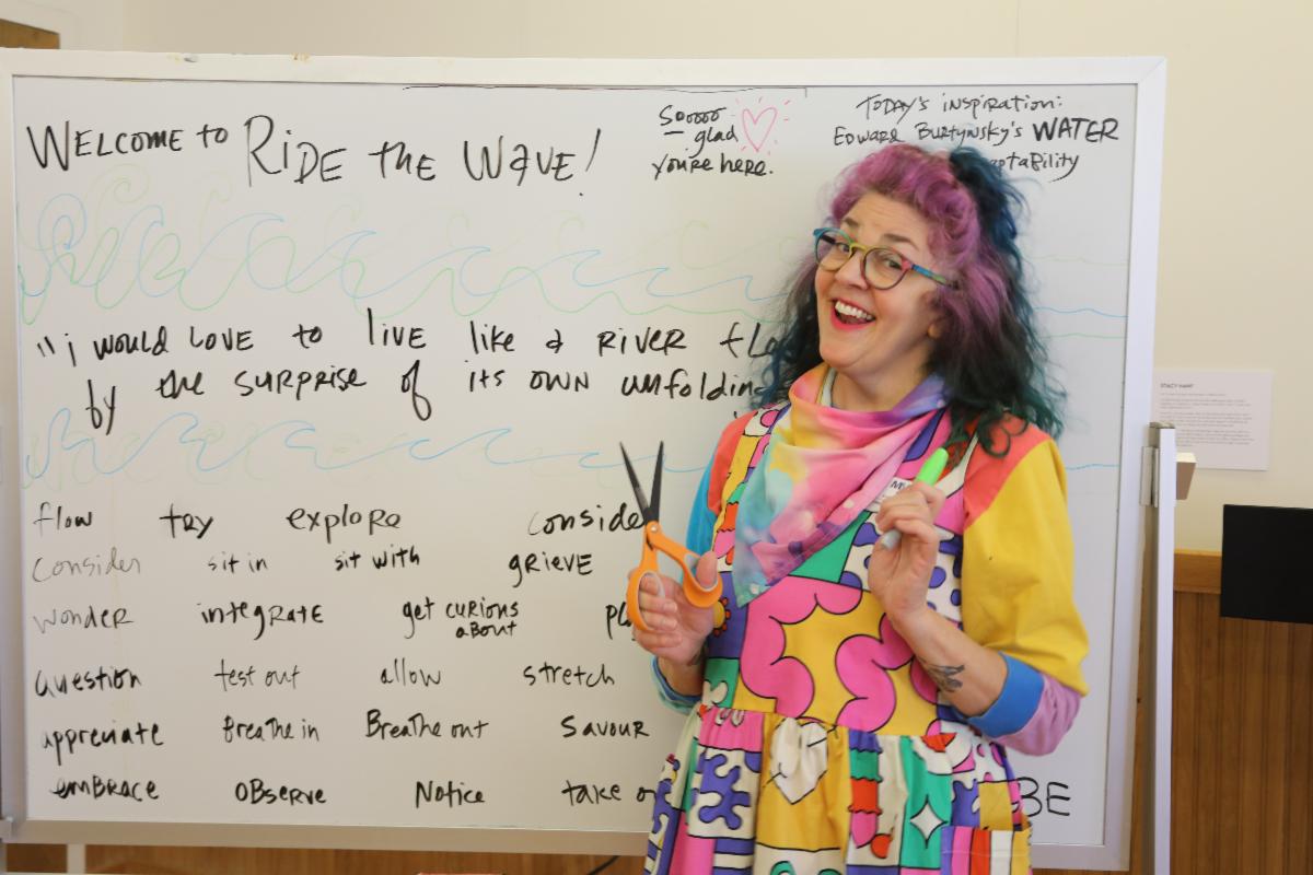 Portrait of program teacher, Sarah Johnson, standing in front of a white board covered with inspirational words, smiling warmly at the camera and holding a pair of scissors. 
